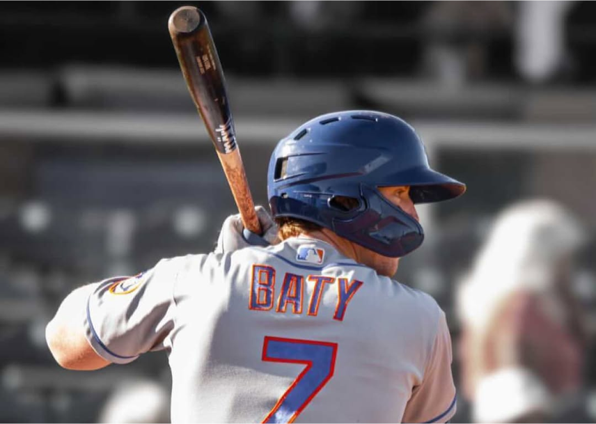 A baseball player wearing a blue helmet and a jersey with the name “Baty” and number 7 prepares to bat, holding a wooden bat over his shoulder.