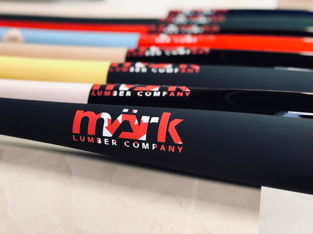 A close-up of several baseball bats featuring the "märk LUMBER COMPANY" logo in red and black, lined up in a row with varying handle colors.