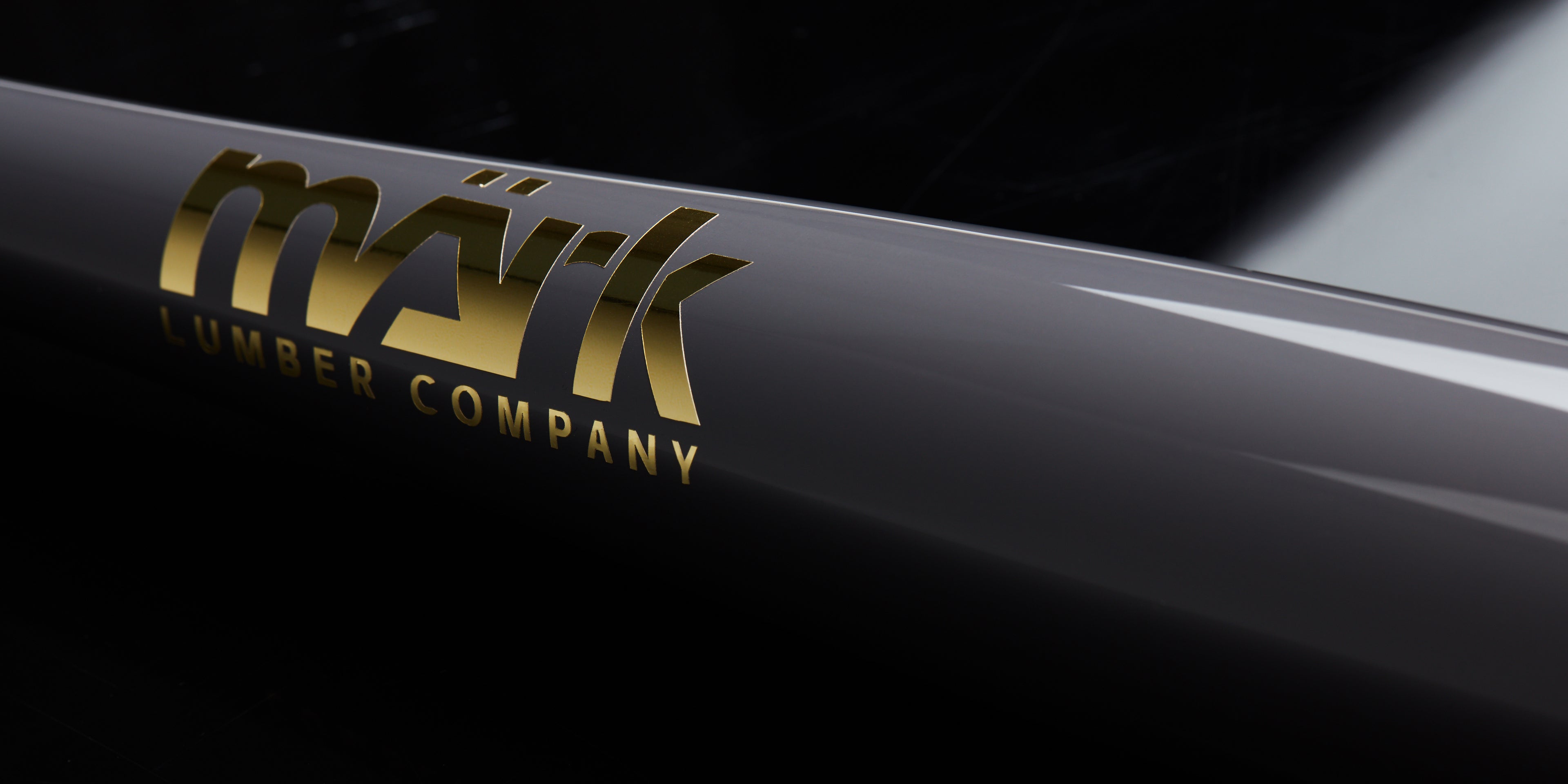Close-up of a glossy black Mark Lumber wood baseball bat featuring gold metallic logo branding, captured against a dark reflective background.