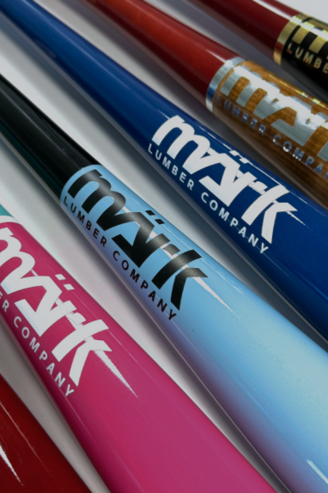 Collection of Mark Lumber wood baseball bats in red, blue, black, pink, and gold finishes with visible logos.