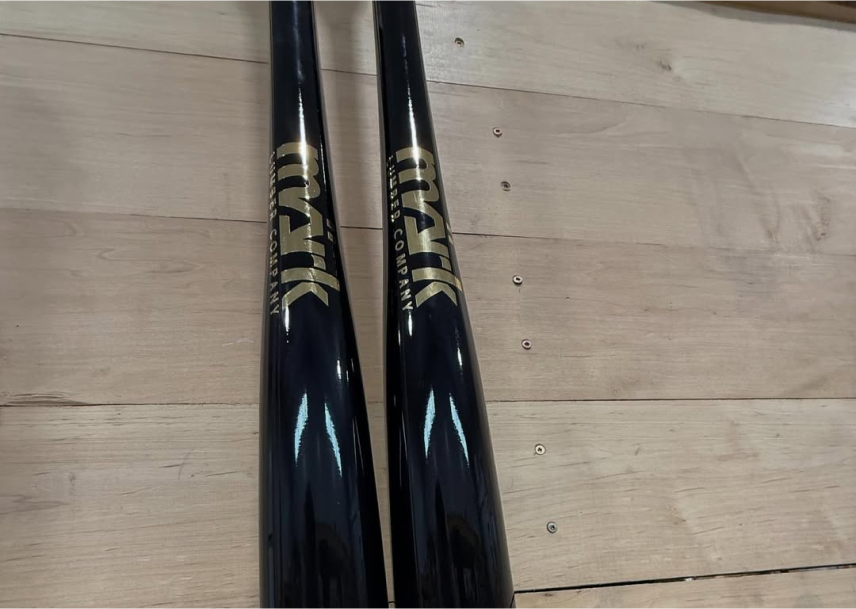 Two glossy black Mark Lumber wood baseball bats with gold logos lying side by side on a wooden surface.