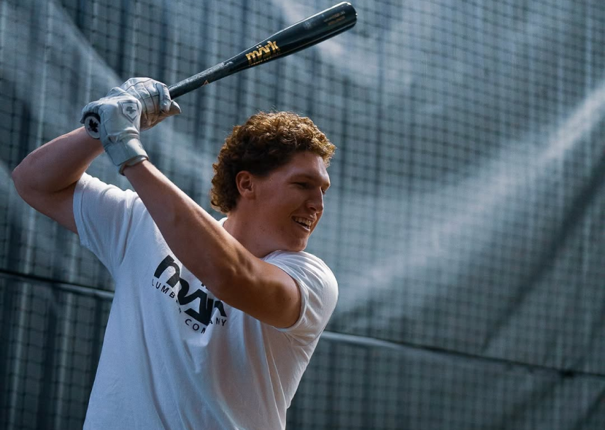 Baseball player swinging a Mark Lumber wood bat while wearing a white Mark Lumber Company t-shirt in a batting cage.