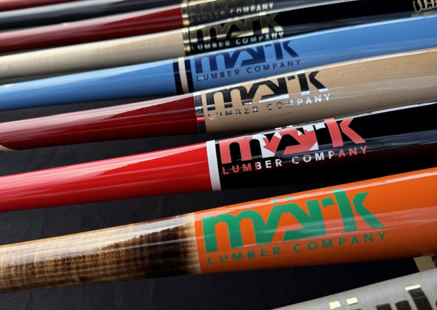 A colorful Mark Lumber wood baseball bats in red, blue, orange, and natural finishes lined up on a black surface.